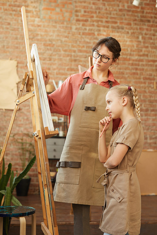 Young teacher standing with little girl near the easel and showing her how to paint a picture during art lesson at studio