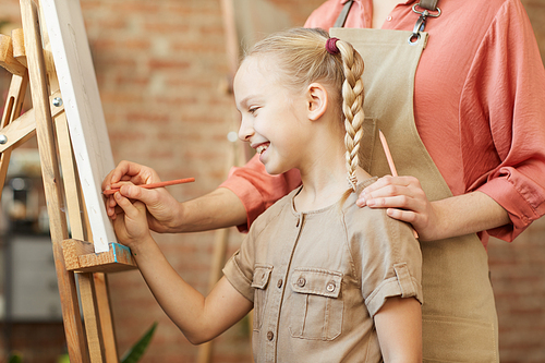 Smiling little girl standing in front of easel and learning to paint with the help of teacher at art studio