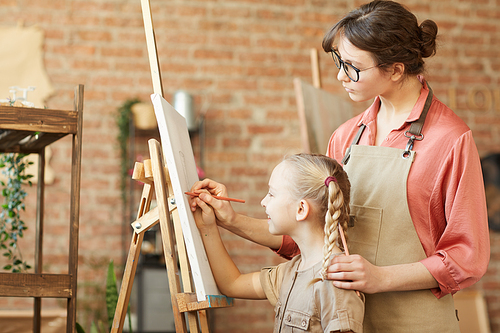 Young teacher helping to paint a picture to little girl while they standing in front of easel at art studio