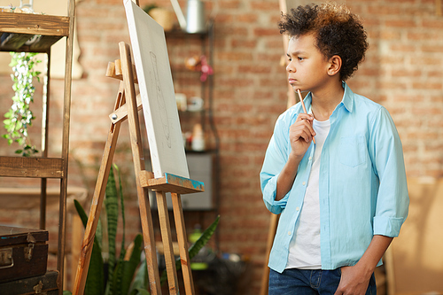 African little boy standing in front of easel and looking at his future picture on it while standing at art studio