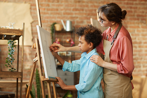 Young teacher standing with African boy near the easel and helping im to paint a picture at art studio