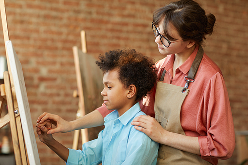 African boy painting on easel with young teacher standing behind him and helping him to paint at art studio