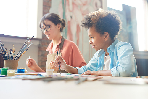 Young woman sitting at the table together with African boy and they painting with watercolors at art studio