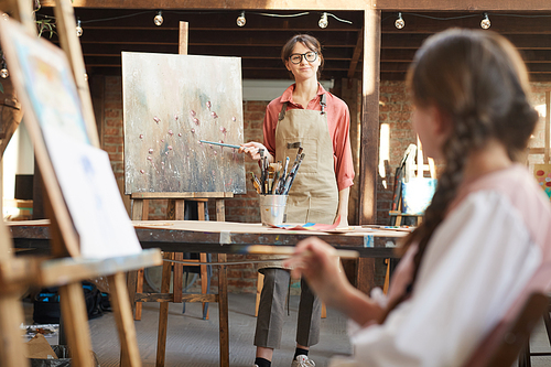 Young teacher standing near the easel pointing at picture and holding art lesson for students at art studio