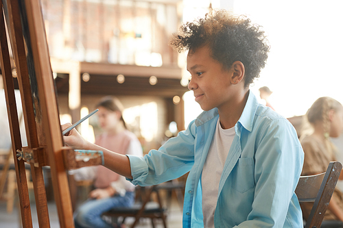 African boy sitting in front of easel and painting a picture on it during art lesson at art studio