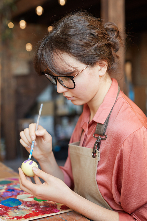 Young woman in eyeglasses sitting at the table and painting an Easter egg for Easter holiday