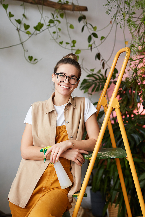 Portrait of young woman in eyeglasses smiling at camera while leaning on the ladder with plants in the background