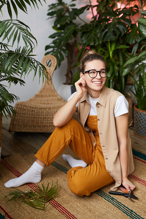 Young happy gardener in eyeglasses sitting on the floor with cut branch and resting