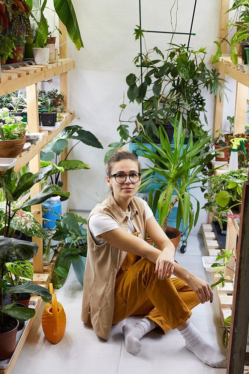 Portrait of young florist in eyeglasses sitting on the floor among green plants and looking at camera in the flower garden