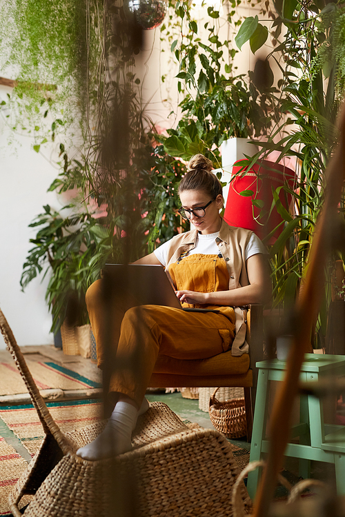 Young woman in eyeglasses sitting on chair and using laptop computer among green plants and flowers