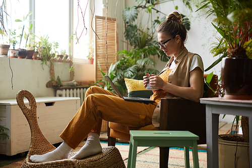 Young woman in eyeglasses sitting on armchair drinking coffee and watching something on laptop in the room with plants