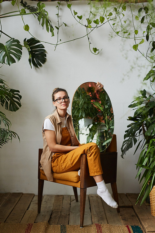 Portrait of beautiful young woman in eyeglasses sitting on armchair and posing at camera with mirror among green plants