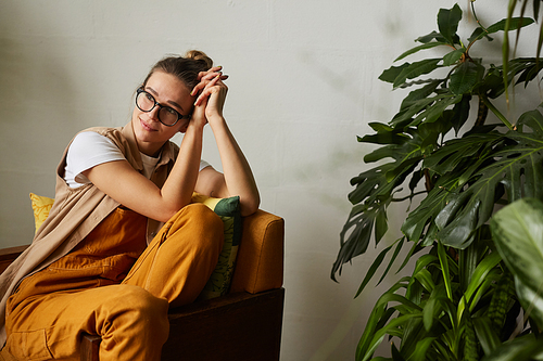 Young woman in eyeglasses resting on armchair in the room with green plants