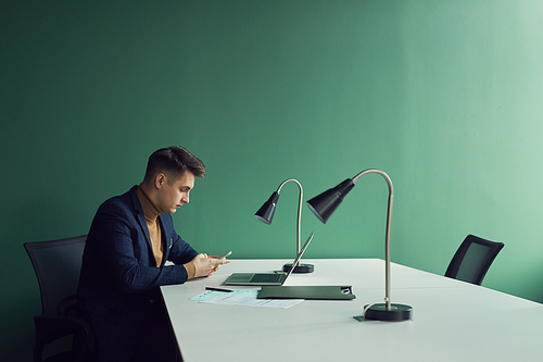 Young serious businessman sitting at his workplace in front of laptop and using mobile phone at office