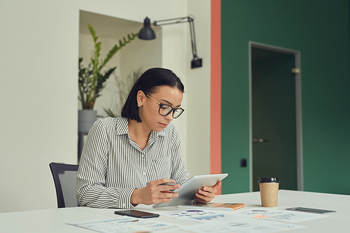 Young serious businesswoman in eyeglasses sitting at her workplace and using digital tablet at office