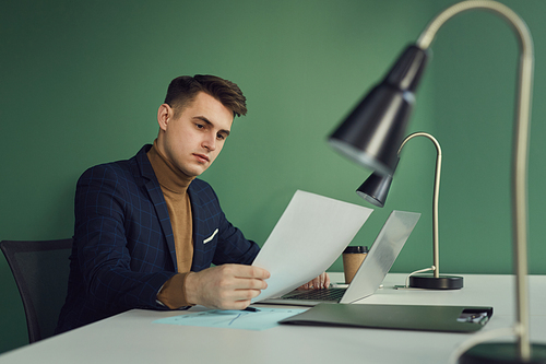 Young businessman sitting at his workplace with laptop computer and examining the document