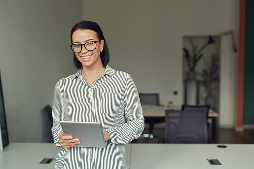 Portrait of young businesswoman in eyeglasses smiling at camera while standing at office with digital tablet