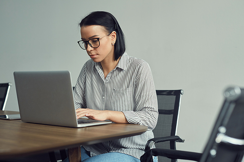 Young businesswoman in eyeglasses concentrating on her online work she sitting at the table and typing on laptop computer