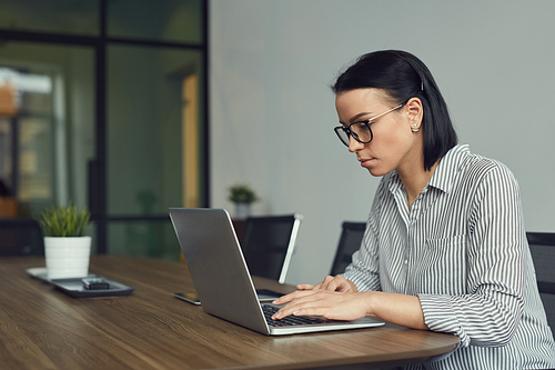 Young office worker in eyeglasses working at her workplace with laptop computer she typing some document or communicating online