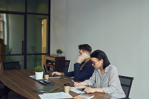 Young businesswoman making notes in document with businessman sitting near be her talking on mobile phone and using laptop they sitting at the table together