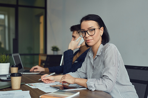 Portrait of young businesswoman in eyeglasses smiling at camera while working at the table with documents with businessman in the background at office