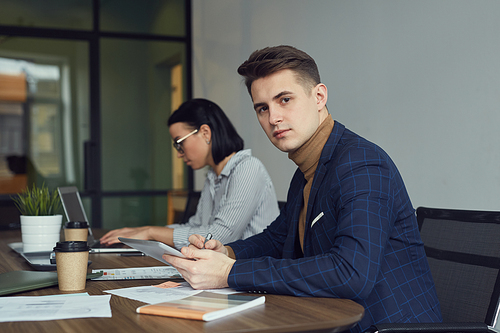 Portrait of young businessman looking at camera and working with tablet pc at the table with businesswoman in the background at office