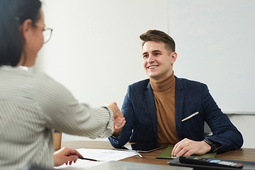 Young smiling businessman sitting at the table and shaking hand to young businesswoman during the meeting at office