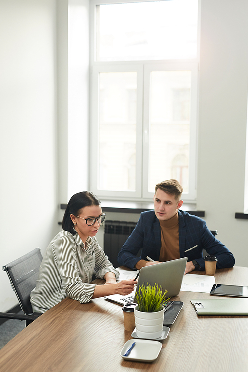 Young businesswoman in eyeglasses pointing at laptop and discussing online presentation together with her colleague while they sitting at the table at office