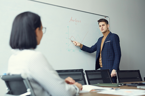 Young businessman standing near the whiteboard and pointing at it he holding presentation fo his colleagues at office