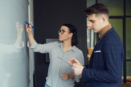 Young businesswoman writing on whiteboard and preparing report together with her colleague before the presentation at office