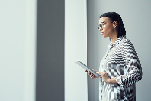 Young serious businesswoman in eyeglasses holding digital tablet and looking through the window while standing at office