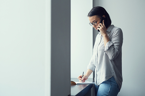 Young businesswoman in eyeglasses standing near the window talking on mobile phone and making note in her notebook