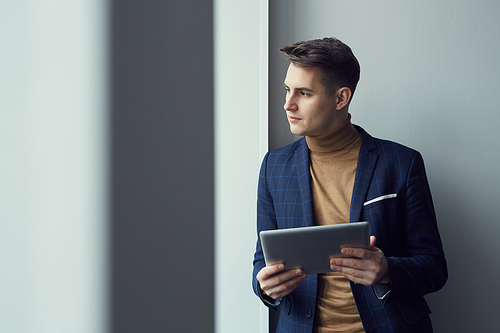 Young businessman in suit standing and looking through the window while using digital tablet