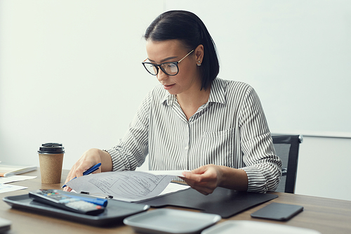 Young businesswoman in eyeglasses sitting at the table with coffee cup and doing paperwork at office