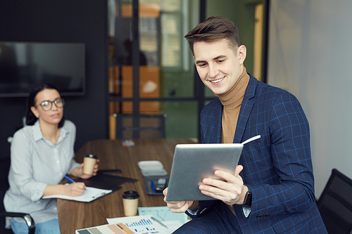 Young businessman holding tablet pc and using it for online communication with businesswoman sitting in the background at office