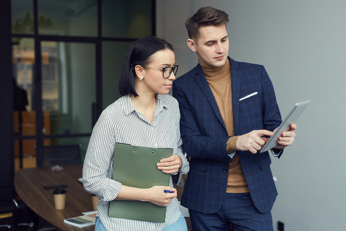 Young businessman pointing at tablet pc and showing something to his colleague while they standing in board room at office