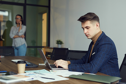 Young businessman sitting at the table typing on laptop computer and working with documents with businesswoman standing in the background at office