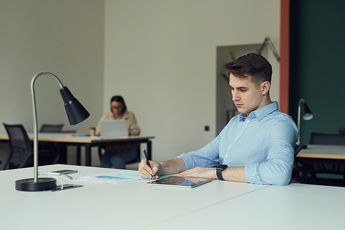 Young businessman sitting at his workplace and doing paperwork with his colleague working in the background at office