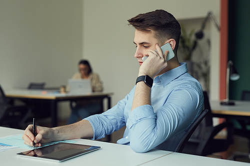 Young businessman talking on mobile phone and making notes in document while working at his workplace at office