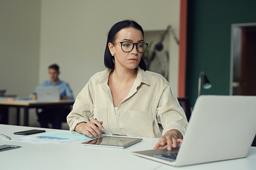 Young serious woman in eyeglasses sitting at the table and using laptop computer in her work at office