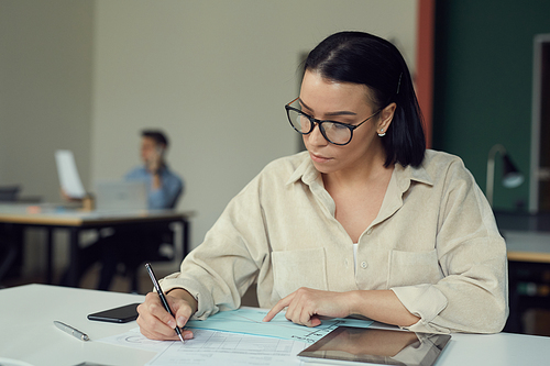 Young manager in eyeglasses sitting at the table and doing paperwork during working day at office