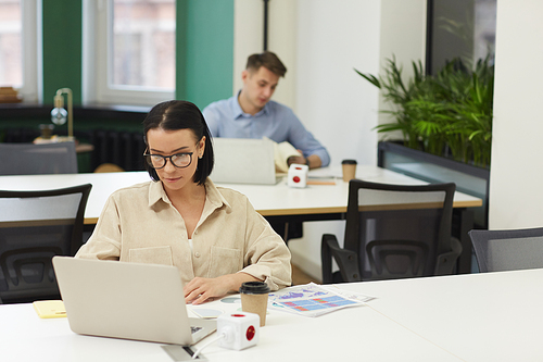 Young businesswoman in eyeglasses sitting at the table and using laptop with other office worker working on laptop in the background