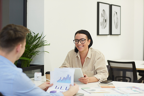 Young businesswoman in eyeglasses smiling to business partner while they discussing financial graphs at the table at meeting