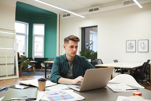 Young businessman sitting at the table among documents and typing on laptop computer he communicating online in modern office