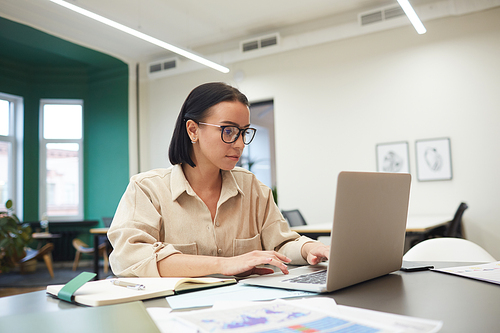 Young businesswoman in eyeglasses sitting at the table and looking at monitor of laptop she doing online work at office