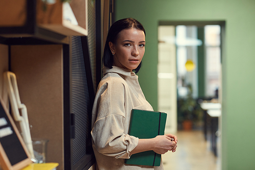 Portrait of young businesswoman holding notepad and looking at camera while standing at office