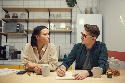 Two young colleagues sitting at the table with notepads and talking to each other during coffee break in the kitchen