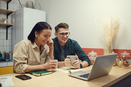 Two business partners sitting at the table drinking coffee and communicating online with their colleague on laptop computer in the kitchen