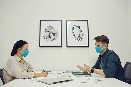 Business partners in protective masks sitting at the table opposite each other and planning work together during meeting at office