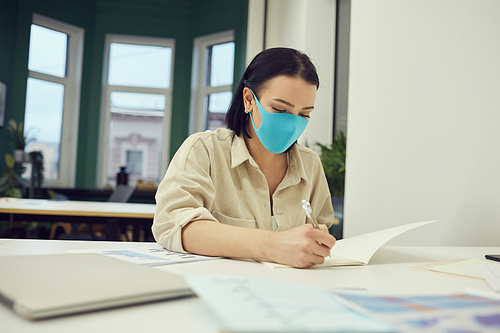 Young businesswoman in protective mask working at her workplace with documents she signing documents at office